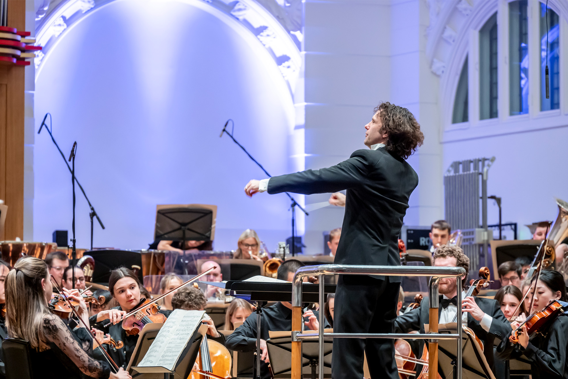 A man, wearing formal attire, conducting an orchestra with his hands, with students performing on their instruments, also wearing formal attire.